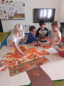 Uczniowie oddziału przedszkolnego stoją przy połączonych stolikach. Dzieci zanurzają dłonie w miseczkach z brązową farbą. Na stolikuułożono tworzoną przez uczniów pracę, gdzie dzieci odciskają swoje dłonie.