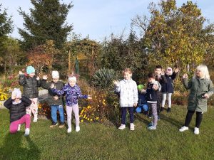 Uczniowie oddziału przedszkolnego stoją w jednym rzędzie wśród roślinności na zielonej polanie. Dzieci machają do zdjęcia.