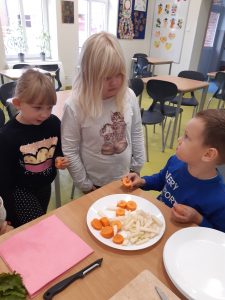 Przy stoliku na szkolnej stołówce siedzichłopiec jedzący warzywa leżące na talerzyku przed nim. Nad chłopcem stoją dwie uczennice oddziału przedszkolnego.