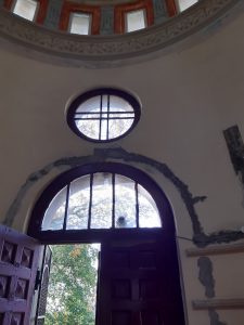 Zdjęcie fragmentu wnętrza kapliczki. Na zdjęciu widoczne są częściowo otwarte drzwi zwieńczone przeszklonym półłukiem oraz okrągłymokienkiem nad nim. Widać ślady prowadzonego w budynku remontu.