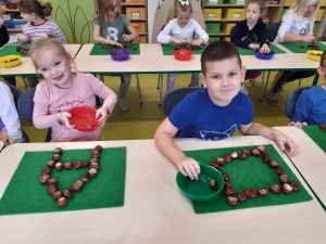 Dwoje uczniów oddziału przedszkolnego siedzi przy wspólnej ławce. Przed nimi na zielonych podkładkach leżą ułożone z kasztanów litery A i kwadrat. Dzieci trzymają miseczki z kasztanami. W tle widać inne dzieci siedzące przy stolikach.
