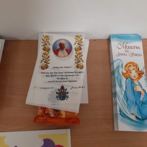 Zdjęcie przygotowanych dla uczestników konkursu nagród. Zakładki z napisem :"MODLITWA DO ANIOŁA STRÓŻA" oraz podobizną ubranego na niebiesko aniołka. Obok leżą białe kartki ze zdjęciem Jana Pawła II oraz fragmentem tekstu. Spod białych kartek wystaje czekolada.