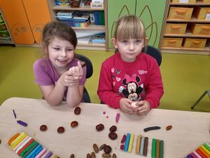 Przy stoliku siedzą dwie uczennice trzymające w dłoniach kasztany. Na stoliku przed nimi leżą kasztany i kolorowe plasteliny.