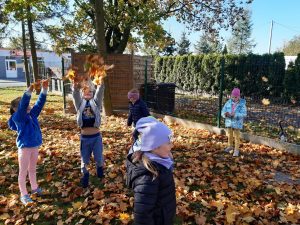 Uczniowie oddziału przedszkolnego chodzą wśród liści na trawniku przy szkolnym budynku. Uczeń stojący po środku podrzuca wysoko w górę liście.