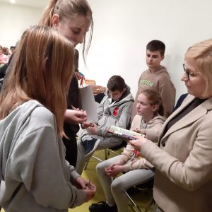 Pani Dyrektor przekazuje nagrodę książkową uczennicom klasy ósmej. W tle widać siedzących na krzesełkach uczniów klasy szóstej.