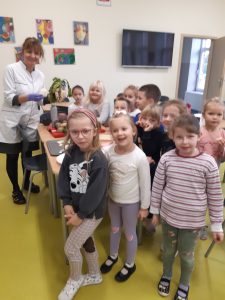 Uczniowie oddziału przedszkolnego stoją w grupie na szkolnej stołówce. Za nimi widać połączone stoliki i Panią nauczycielkę ubraną w biały kitel i trzymającą w dłoniach warzywo z natką.