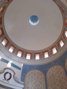 Zdjęcie okrągłego sufitu kapliczki z prostokątnymiokienkami ustawionym w okręgu. Na zdjęciu widać fragmenty ścian zdobionych na brązowo - niebiesko.