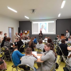 Uczniowie siedzą przy stolikach podczas konkursu wiedzy o Janie Pawle II. Przed nimi na tle zdjęcia Jana Pawła II stoi Pani katechetka trzymająca złożone przed sobą dłonie.