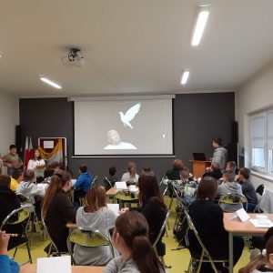 Uczniowie siedzą przy stolikach podczas konkursu wiedzy o Janie Pawle II. Na ścianie przed nimi wyświetla się zdjęcie Jana Pawła II do którego nadlatuje biały gołąbek.