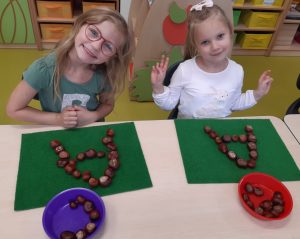 Dwie uczennice oddziału przedszkolnego siedzą przy wspólnej ławce. Przed nimi na zielonych podkładkach leżą ułożone z kasztanów litery A, a przed nimi stoją miseczki z kasztanami.