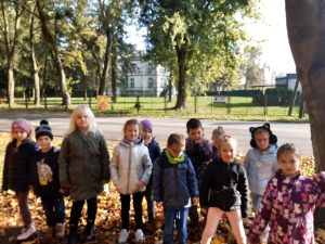 Uczniowie oddziału przedszkolnego stoją obok siebie na trawniku przykrytym liśćmi. Za nimi widać drogę oraz ukryty wśród drzew pałacyk.