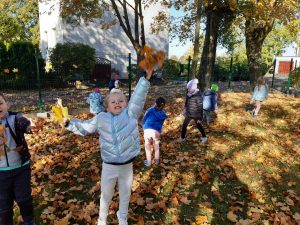 Uczniowie oddziłu przedszkolnego chodzą wśród liści na placu przy szkole. Dziewczynka stojąca na pierwszym planie podrzuca w górę zgromadzone w dłoniach liście.
