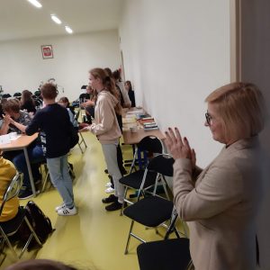 Pani Dyrektor stojąc bokiem do aparatu klaszcze. W tle widać również stojących bokiem uczniów klasy szóstej.