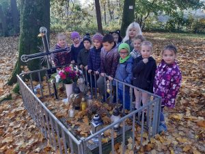 Uczniowie oddziału przedszkolnego stoją obok siebie wzdłuż niskiego ogrodzenia nagrobku z krzyze. W okół dzieci i na nagrobku leżą liście.