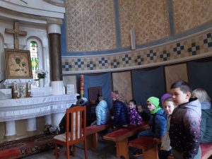 Uczniowie siedzą na ławeczkach wewnątrz kapliczki. Dzieci zwrócone są twarzą w stronę ołtarzu z ustawionym obrazem Matki Bożej oraz zawieszonym nad nim krzyżem. Nad dziećmi widoczne są fragmenty brązowo-niebieskiej mozaiki i brązowe zdobienia ścian.