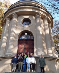 Uczniowie oddziału przedszkolnego stoją w grupie w wejściu do kapliczki. Na zdjęciu widoczny jest cały budynek okragłej kapliczki z kolumnami.