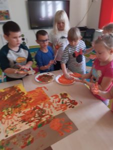 Uczniowie oddziału przedszkolnego stoją przy połączonych stolikach. Dzieci zanurzają dłonie w miseczkach z brązową farbą. Na stolikuułożono tworzoną przez uczniów pracę, gdzie dzieci odciskają swoje dłonie.