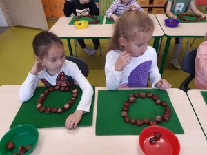 Dwie uczennice oddziału przedszkolnego siedzą przy wspólnej ławce. Przed nimi na zielonych podkładkach leżą ułożone z kasztanów kółeczka, a przed nimi stoją miseczki z kasztanami. Dziewczynki patrzą w bok a za nimi widać inne dzieci siedzące przy stolikach.