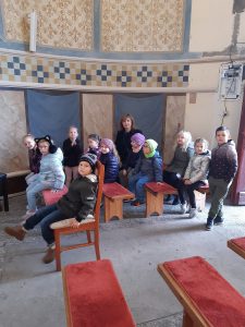 Uczniowie oddziału przedszkolnego wraz z swoją Panią siedzą na ławeczkach wewnątrz kapliczki. Jeden chłopiec stoi. Nad dziecmi widać brązowo - niebiesko zdobione ściany kapliczki.