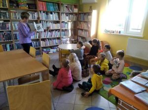 Pani Monika trzyma w dłoniach otwarta książkę, którą pokazuje siedzącym na podłodze uczniom. Zdjęcie wykonane w gminnej bibliotece.