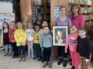 Uczniowie oddziału przedszkolnego pozuja do zdjęcia z Panią Moniką i swoją nauczycielką. Pani Monika trzyma w dłoniach otrzymany prezent - pracę plastyczną jednej z uczennic naszej szkoły. Zdjęcie wykonane w bibliotece w Ostaszewie.