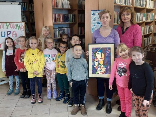 Uczniowie oddziału przedszkolnego pozuja do zdjęcia z Panią Moniką i swoją nauczycielką. Pani Monika trzyma w dłoniach otrzymany prezent - pracę plastyczną jednej z uczennic naszej szkoły. Zdjęcie wykonane w bibliotece w Ostaszewie.