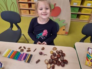 Uśmiechnięta uczennica siedzi za stolikiem, na którym leżą kasztany i kolorowe plasteliny w pudełku.