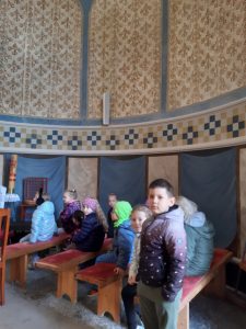 Uczniowie oddziału przedszkolnego siedzą na ławeczkach wewnątrz kapliczki. Nad głowami dzieci widać brązo - niebieskie zdobione ściany kapliczki.