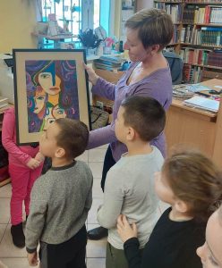 Pani Monika podziwia otrzymany prezent - oprawioną w ramkę pracę plastycznąjednej z uczennic. Obok niej stoją uczniowie oddziału przedszkolnego. Zdjęcie wykonane w bibliotece w Ostaszewie.