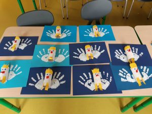 Zdjęcie ułozonych na stoliku prac uczniów z oddziału przedszkolnego. Dzieci na niebieskich kartkach zrobiły białe odciski swoich rąk a pomiędzy nie wkleiły wukonane z białych rolek i ozdobione na żóło podobizny orłów.