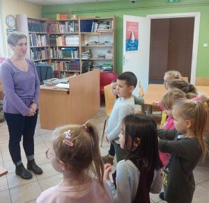 Pani Monika stoi na przeciwko uczniów oddziału przedszkolnego i do nich przemawia. Zdjęcie wykonane w bibliotece w Ostaszewie.