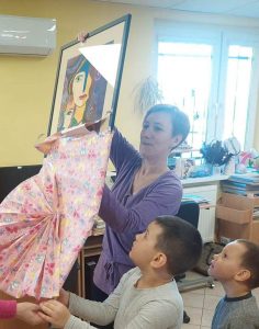 Pani Monika rozpakowuje otrzymany prezent. Pomafają jej w tym ucznioiwe oddziału przedszkolnego. Dzieci trzymają papier, w który zapakowany był pezent.