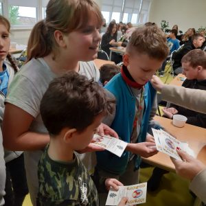 Troje uczniów klasy czwartej stoi obok siebie trzymając w rękach kolorowe, nadrukowane karteczki. Z boku widać dłonie Pani Dyrektor z kolejnymi karteczkami. W tle widać siedzących przy stolikach innych uczniów.