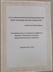 Zdjęcie dedykacji przygotowanej dla Pani Moniki. Na kremowym tle cytat : "To za sprawą bezinteresownego poświęcenia ludzie nawiązują łączność między sobą" Antoine de Saint-Exupery. Poniżej dedykacja: "W podziękowaniu za wieloletnią współpracę Dyrektor, Pracownicy i Uczniowie Szkoły Podstawowej w Ostaszewie". U dołu zamieszczono datę "Ostaszewo 08.11.2022".