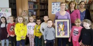 Pani Monika pozuje do zdjęcia razem z uczniami oddziału przedszkolnego, ich nauczycielką oraz Panią Sylwią - szkolną bibliotekarką. Pani Monika trzyma w dłoniach otrzymany prezent - pracę plastyczną jednej z uczennic naszej szkoły.