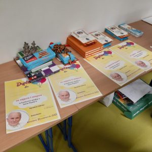 Zdjęcie przygotowanych w konkursie nagród. Na stoliku leżą cztery dyplomy ze zdjęciem Jana Pawła II a nad nimi, ułożone w kupki czekolady, notesy i różańce.