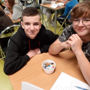 Dwóch uczniów klasy ósmej siedzi przy wspólnym stoliku, na którym stoi miseczka z cukierkami. Chłopcy szeroko uśmiechają się do zdjęcia.