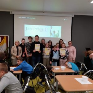 Uczniowie wraz z Panią Dyrektor oraz dwie nauczycielkami pozują do zdjęcia grupowego. Troje uczniów trzyma przed sobą dyplomy. Wszyscy stoją na tle ściany, na której wyświetla się napis JAN PAWEŁ II oraz podobizna papieża. Przed fotografowaną grupą przy stolikach siedzą pozostali uczniowie.