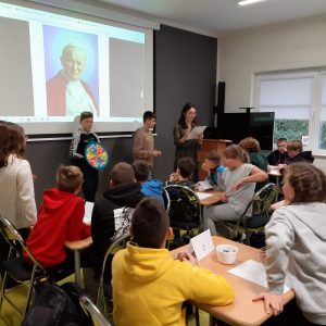 Uczniowie siedzą przy stolikach podczas konkursu wiedzy o Janie Pawle II. Przed nimi na ścianie wyświetla się portret Jana Pawła II na tle którego stoi uczeń z kółkiem podzielonym za pomocą kolorów na wycinki. Obok ucznia stoi Pani nauczycielka z kartką.