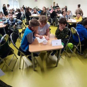 Uczniowie siedzą przy stolikach podczas konkursu wiedzy o Janie Pawle II. Dzieci rozwiązują na kartkach zadania.