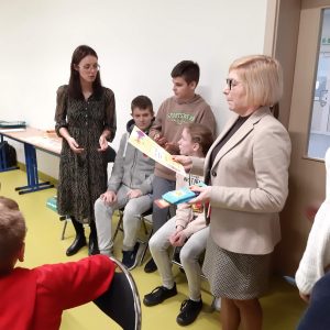 Pani Dyrektor trzyma w jednej dłoni dyplom a w drugiej czekoladę. Obok niej siedzi na krzesełkach trójka uczniów klasy szóstej a dalej stoi Pani nauczycielka.