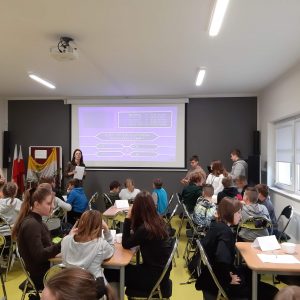 Uczniowie siedzą przy stolikach podczas konkursu wiedzy o Janie Pawle II. Przed nimi na ścianie wyświetla się guiz z pytaniem w formie jak z milionerów. Obok guizu stoi Pani nauczycielka z białą kartką w dłoniach.