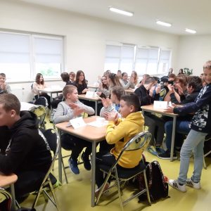 Uczniowie siedzą przy stolikachpodczas konkursu wiedzy o Janie Pawle II. Dzieci klaszczą, a na stolikach mają ustawione białe kartki.