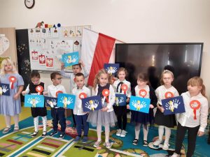 Uczniowie oddziału przedszkolnego w strojach galowych pozują do wspólnego zdjęcia. Dzieci maja przyczepiono kotyliony a w rękach trzymają wykonane przez siebie prace przedstawiające orły. W tle za nimi zza telewizora wystaje bokiem flaga Polski.