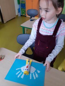 Uczennica oddziału przedszkolnego stoi przed stolikiem o który opiera wykonaną przez siebie pracę przedstawiającą orła w koronie. Na niebieskim tle odcisnęła w białym kolorze swoje dłonie a pomiędzy przykleiła orła wykonanego z bałej rolki z koroną i żółtymi nogami.