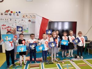 Uczniowie oddziału przedszkolnego w strojach galowych pozują do wspólnego zdjęcia. Dzieci maja przyczepiono kotyliony a w rękach trzymają wykonane przez siebie prace przedstawiające orły. W tle za nimi zza telewizora wystaje bokiem flaga Polski.