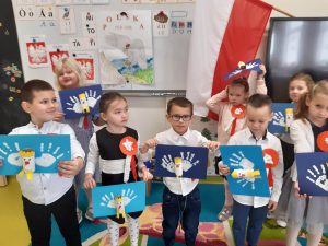 Uczniowie oddziału przedszkolnego w strojach galowych pozują do wspólnego zdjęcia. Dzieci maja przyczepiono kotyliony a w rękach trzymają wykonane przez siebie prace przedstawiające orły. Za nimi widać zawieszoną flagę Polski.