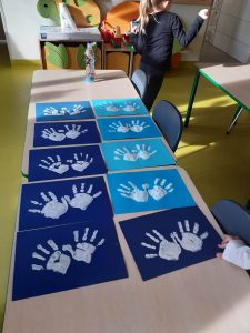 Zdjęcie ułożonych w dwóch rzędach na stoliku prac uczniów, jeszcze przed ostatecznym dokończeniem. Na niebieskim tle dzieci odcisneły na biało swoje rozpostarte dłonie.