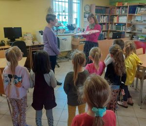 Pani Alicja - wychowawczyni oddziału przedszkolnego, wręcza Pani Monice prezent przygotowany z okazji jej odejścia z biblioteki w Ostaszewie. Scenie przyglądają się uczniowie oddziału przedszkolnego.