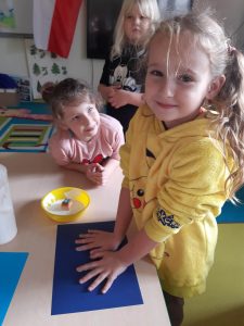 Uczennica podczas robienia odcisku dloni na niebieskiej kartce. Dziewczynka mocno przyciska swoje dlonie do kartki leżącej na stoliku. Obok z opartymi na stoliku przedrabieniami stoi koleżanka przypatrująca się jej z zaciekawieniem.